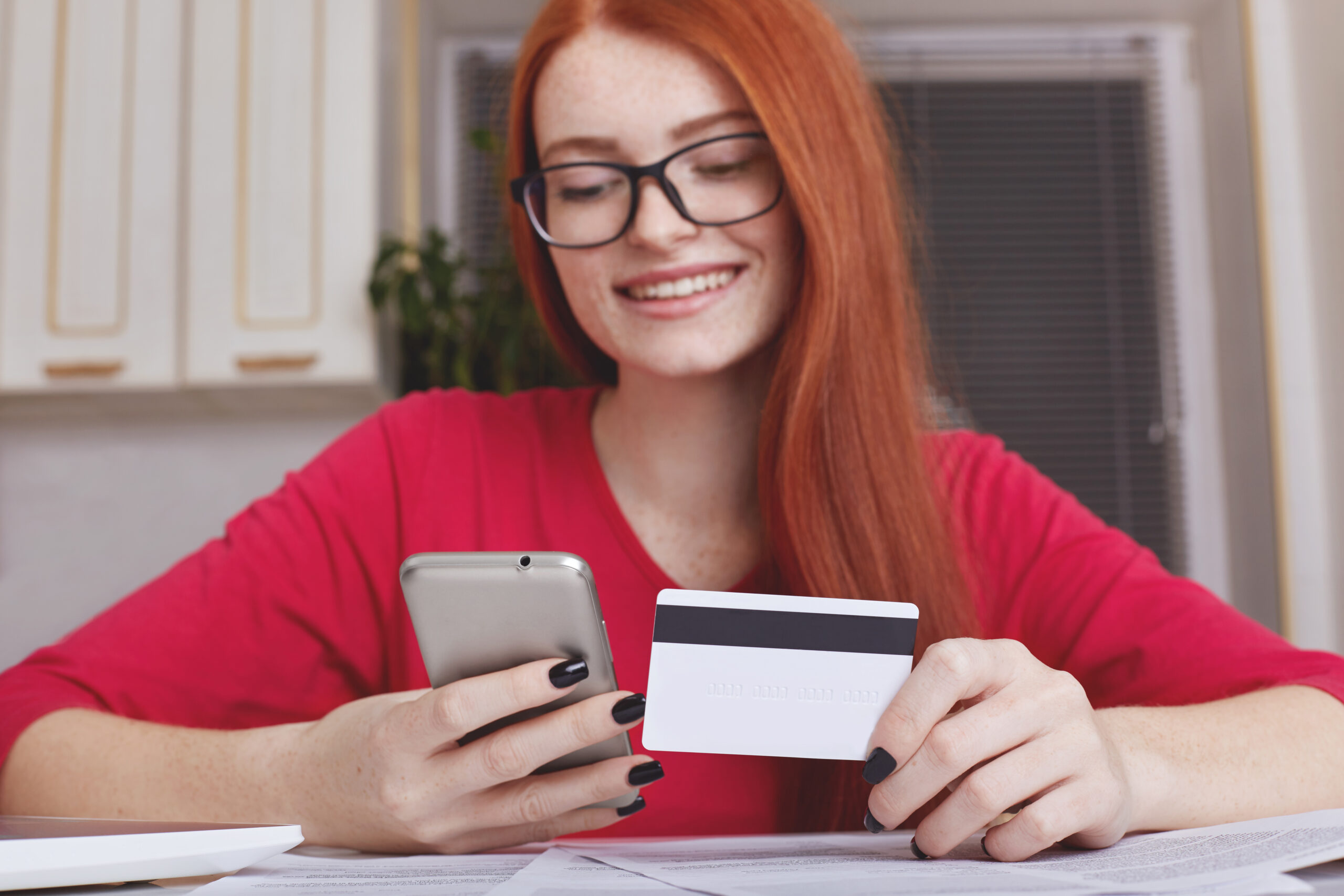 Redhaired pretty female model in eyewear holds smart phone and credit card, makes online purchase or shopping in internet store, has happy expression. Online payment. Mobile payments concept